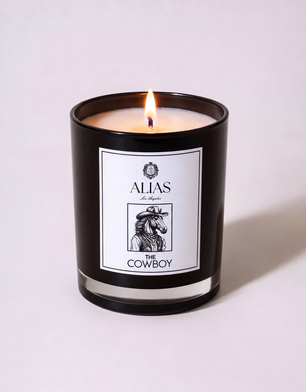 A lit black glass jar candle with cream wax on a light gray background. The front label reads 'ALIAS' and 'THE COWBOY' with a black-and-white cowboy illustration.
