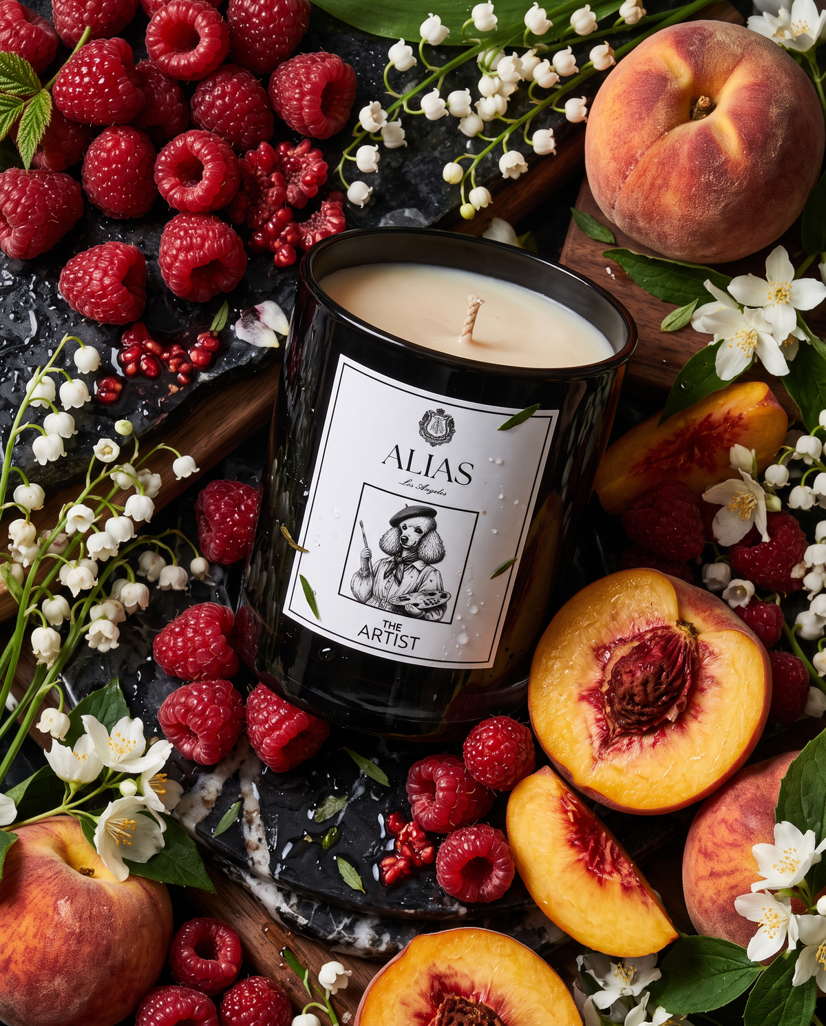 A black glass candle jar labeled 'ALIAS for ARTIST' is styled in a flat lay with fresh raspberries, peaches, white flowers, and green leaves on a dark stone surface.