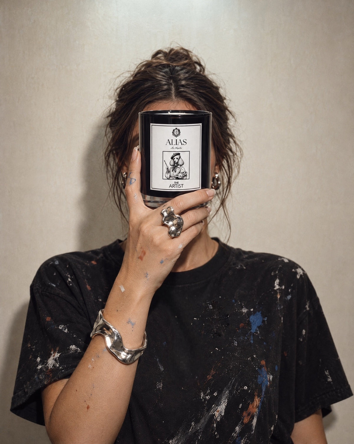 A person in a paint-splattered black t-shirt holds a black glass jar candle in front of their face. The candle label reads 'ALIAS LOS ANGELES' and 'THE ARTIST'.