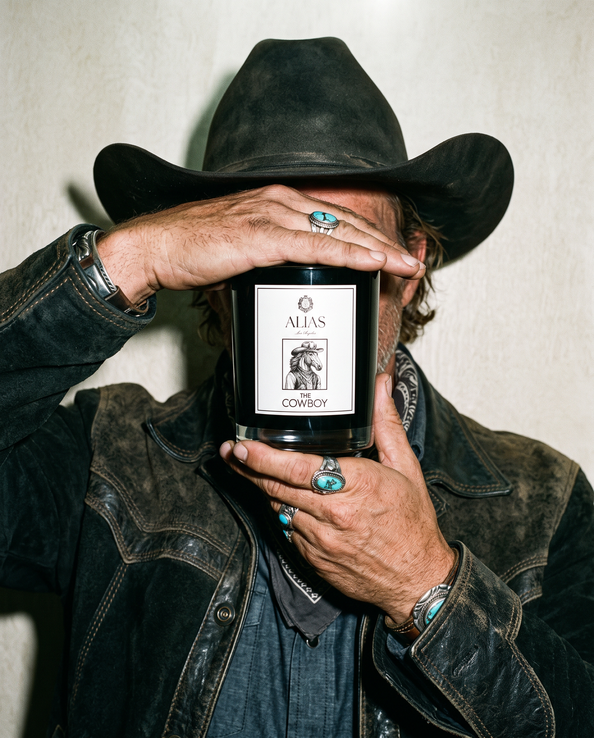 A man in a black cowboy hat and distressed leather jacket holds a black candle with a white label that reads 'ALIAS COWBOY' in front of his face. He is wearing silver rings with turquoise stones and a silver watch, against a plain off-white wall.