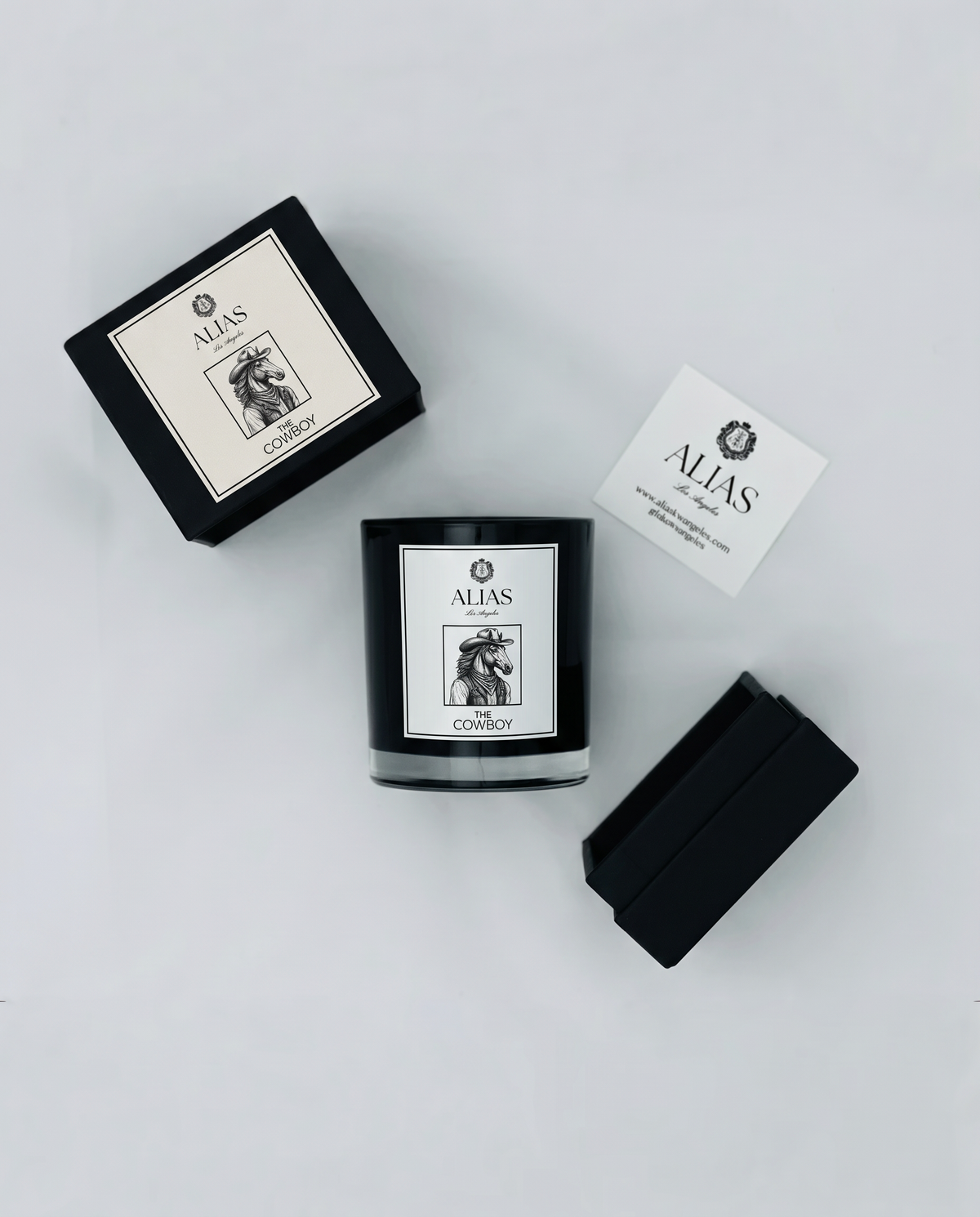 A black glass candle jar labeled 'ALIAS LOS ANGELES THE COWBOY' is arranged on a light gray background with its black box, a small white card, and another black box piece. The visible brand is Alias.